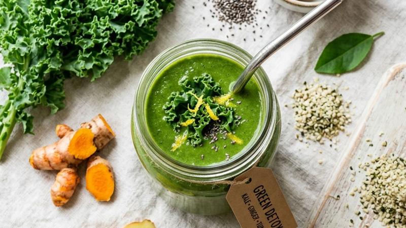 A tall glass of bright green smoothie with a straw, fresh ginger, and lemon slices on a wooden cutting board